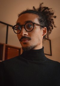Homme aux cheveux bouclés attachés, portant des lunettes rondes noires et un col roulé noir, regardant sur le côté en intérieur.