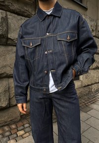 Τζάκετ τζιν σε σκούρο μπλε χρώμα, με δύο τσέπες στο στήθος, χάλκινους κουμπιά και αντίθετη κίτρινη ραφή. Συνδυάζεται με αντίστοιχα τζιν παντελόνια.