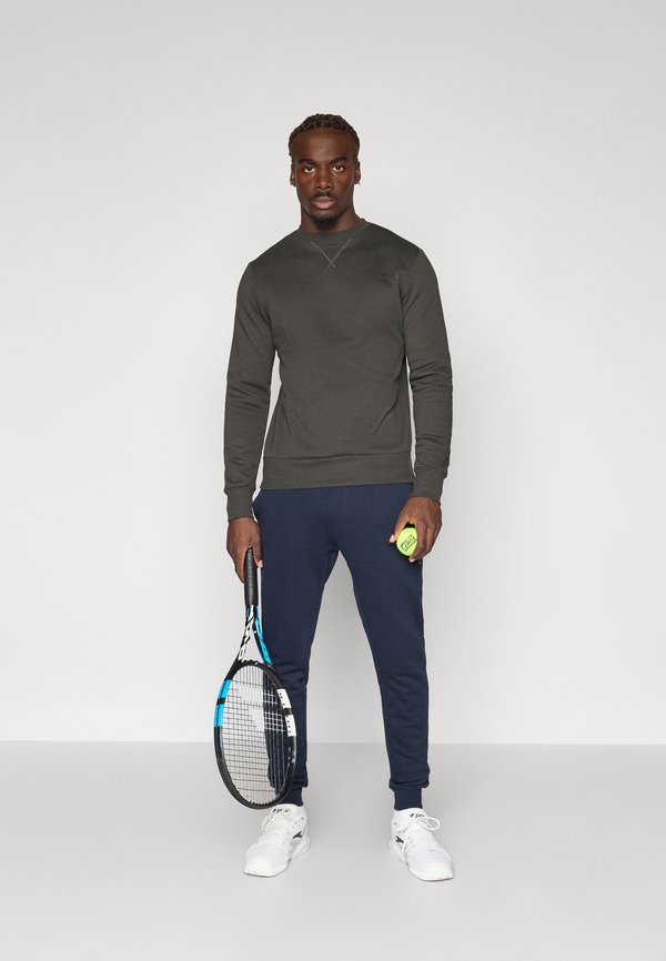 Sudadera verde oscuro con cuello redondo, combinada con pantalones jogger azul marino. Sosteniendo una raqueta de tenis y una pelota de tenis amarilla brillante.