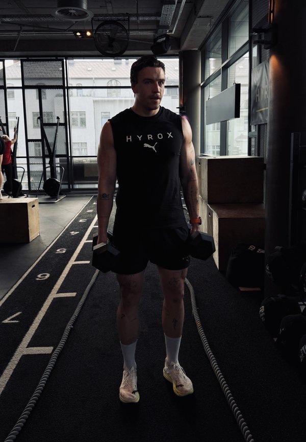 Homme en débardeur HYROX se tient à l’intérieur, tenant des haltères sur les côtés, sur une piste numérotée avec du matériel de gym et des caisses à proximité.