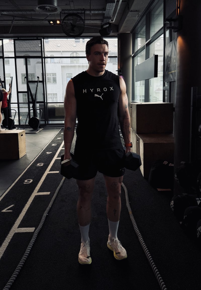 Homme en débardeur HYROX se tient à l’intérieur, tenant des haltères sur les côtés, sur une piste numérotée avec du matériel de gym et des caisses à proximité.