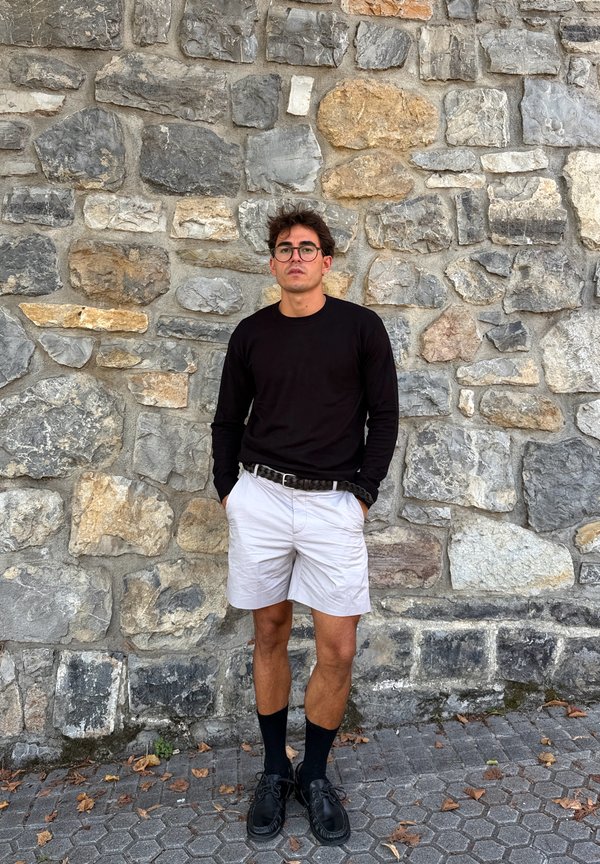 Ung mand med briller iført sort langærmet skjorte, hvide shorts, sorte sokker og sko, står op ad en tekstureret stenvæg.