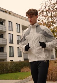 Veste de sport blanche avec des accents gris, leggings noirs avec logo, et gants noirs. Personne courant le long d'un chemin près de bâtiments modernes.