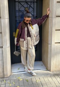 Femme portant une veste matelassée à blocs de couleurs, un pantalon ample crème et des baskets, debout contre une porte encadrée de pierre avec des barreaux noirs, tenant un sac à main.