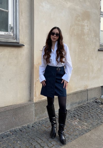 Camisa blanca de manga larga, falda envolvente de mezclilla oscura con dobladillo asimétrico, botas negras hasta la rodilla, de pie contra una pared texturizada.