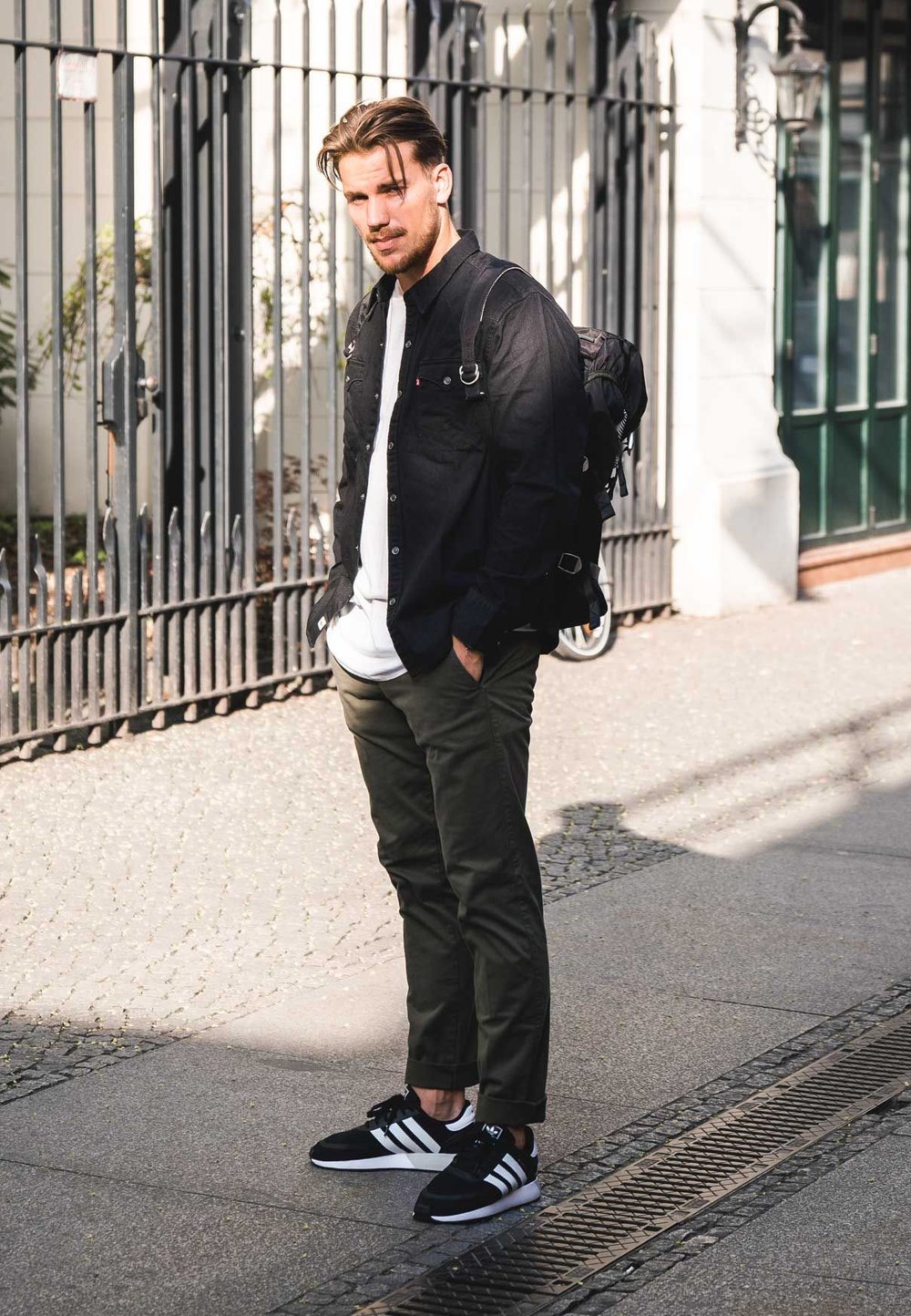 Schwarzes Hemd mit Knöpfen, weißes T-Shirt, olivgrüne Hose, schwarz-weiße Sneaker. Rucksack mit sichtbaren Riemen auf dem Rücken.