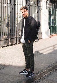 Schwarzes Hemd mit Knöpfen, weißes T-Shirt, olivgrüne Hose, schwarz-weiße Sneaker. Rucksack mit sichtbaren Riemen auf dem Rücken.