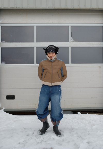 Joven con chaqueta marrón, vaqueros holgados, botas negras y orejeras, de pie sobre la nieve frente a una puerta de garaje blanca.