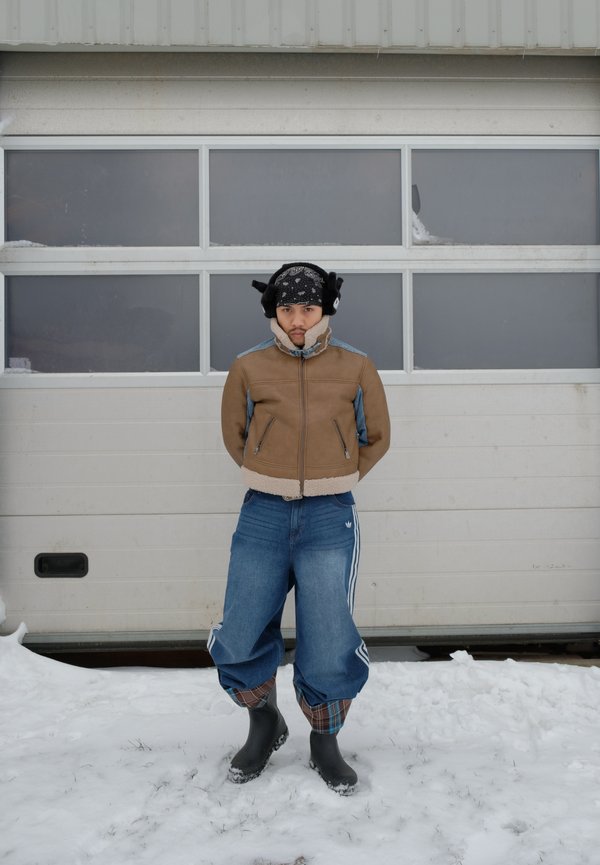 Junge Person trägt eine braune Jacke, weite Jeans, schwarze Stiefel und Ohrenschützer, steht im Schnee vor einer weißen Garagentür.