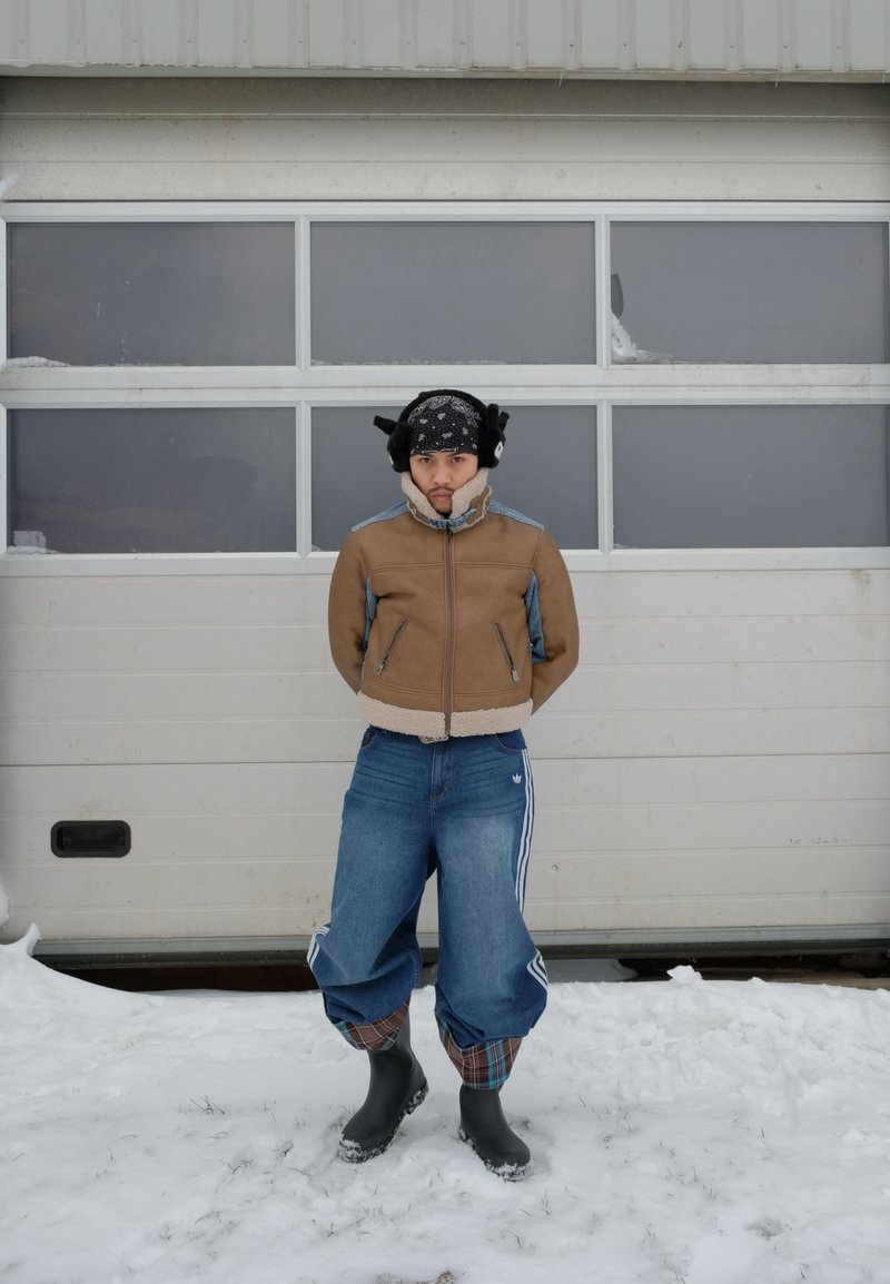 Ung person som bär en brun jacka, lösa jeans, svarta stövlar och öronmuffar står på snö framför en vit garageport.