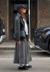 Czarna skórzana kurtka, biały sweter, szara maxi spódnica w paski, białe skarpetki, czarne buty, czapka, trzyma torbę z zaokrąglonym uchwytem.