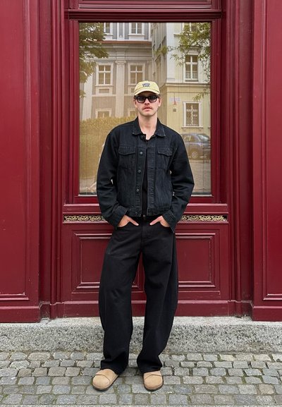 Chaqueta de denim negra con dos bolsillos frontales, pantalones negros, zapatos beige de tipo deslizante y una gorra de béisbol amarillo claro, contra una pared roja.