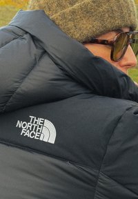 Άτομο που φοράει μαύρο μπουφάν The North Face, καφέ πλεκτό σκουφί και γυαλιά ηλίου με σχέδιο χελώνας, κοιτάζοντας προς τα πλάγια σε εξωτερικό χώρο.