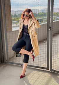 Beige Trenchcoat mit doppelter Knopfreihe, schwarzes Spitzentop, eng sitzende schwarze Cropped-Hosen und burgunderrote Spitzer-Pumps. Gitter-Hintergrund.