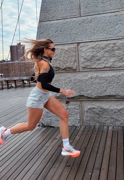 Corredor con camiseta de manga larga negra, pantalones cortos grises y zapatillas blancas con detalles neón, corriendo sobre una superficie de madera cerca de una pared de piedra.