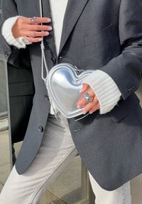 Händer som håller en silverfärgad hjärtformad väska, bärandes en mörkgrå kavaj, en vit tröja med långa ärmar, ljusa jeans och silverringar.