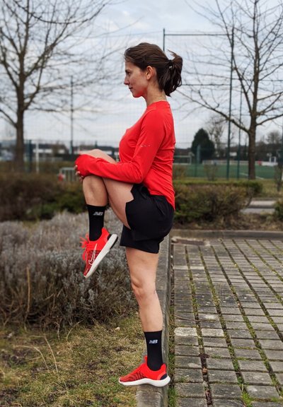 Camiseta atlética roja de manga larga, pantalones cortos negros, calcetas negras de New Balance y zapatillas rojas. Persona estirándose individualmente con la rodilla levantada en un entorno al aire libre.