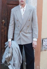 Lysgrå dobbeltbrystet blazer med lodrette striber, knapper og to forlommer; model holder en matchende lys jakke; mørke bukser.