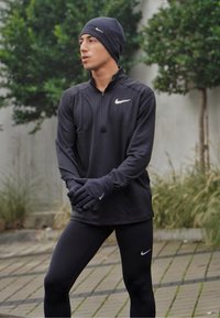 Fekete Nike zippzáras póló hosszú ujjú, fekete leggings-szel és kesztyűvel párosítva. Az pólón és a kesztyűn ezüst Nike logó található a mellkason.