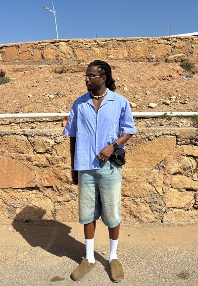 Camisa de manga corta a rayas azul claro, pantalones cortos de mezclilla desgastados, calcetines blancos, zapatos de eslora marrones, bolso cruzado negro, entorno al aire libre.