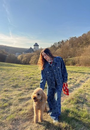 Denim-Outfit mit floralem Muster, übergroßer Jacke und weit geschnittenen Hosen in Blau. Ein hellbrauner Hund mit lockigem Fell steht daneben.