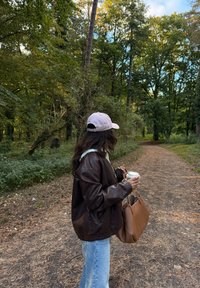 Veste en cuir marron, jean bleu clair, hoodie gris clair, sac à main marron, tasse de café blanche, à l'extérieur sur un chemin boisé avec des arbres.