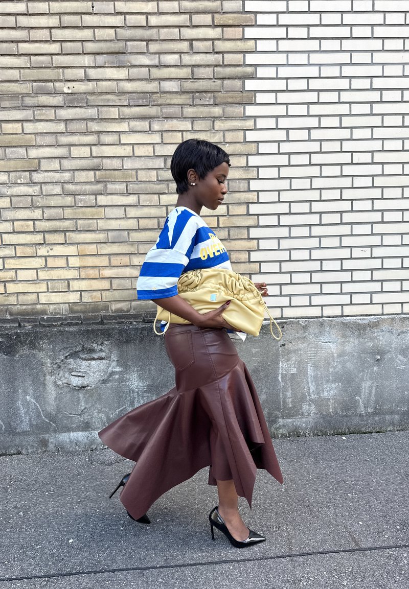 Tunique oversize rayée bleue et blanche, jupe asymétrique en similicuir marron, escarpins noirs à bout pointu et sac à main jaune plissé.