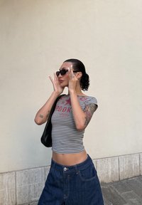 Raidallinen harmaa crop-top, jossa on punaisia kirjaimia, farkkuhame, mustat aurinkolasit ja musta laukku. Huomiota herättäviä tatuointeja käsivarsissa, keskipitkät kiharat hiukset.