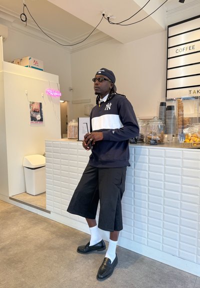 Sudadera negra y blanca con el logo de NY, pantalones cortos negros, calcetines blancos y mocasines negros. El interior presenta un mostrador de azulejos blancos y un letrero de neón rosa.