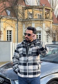 Un homme portant une veste à carreaux noir et blanc et des lunettes de soleil se tient près d'une voiture Audi argentée devant une maison jaune avec des tuiles rouges.