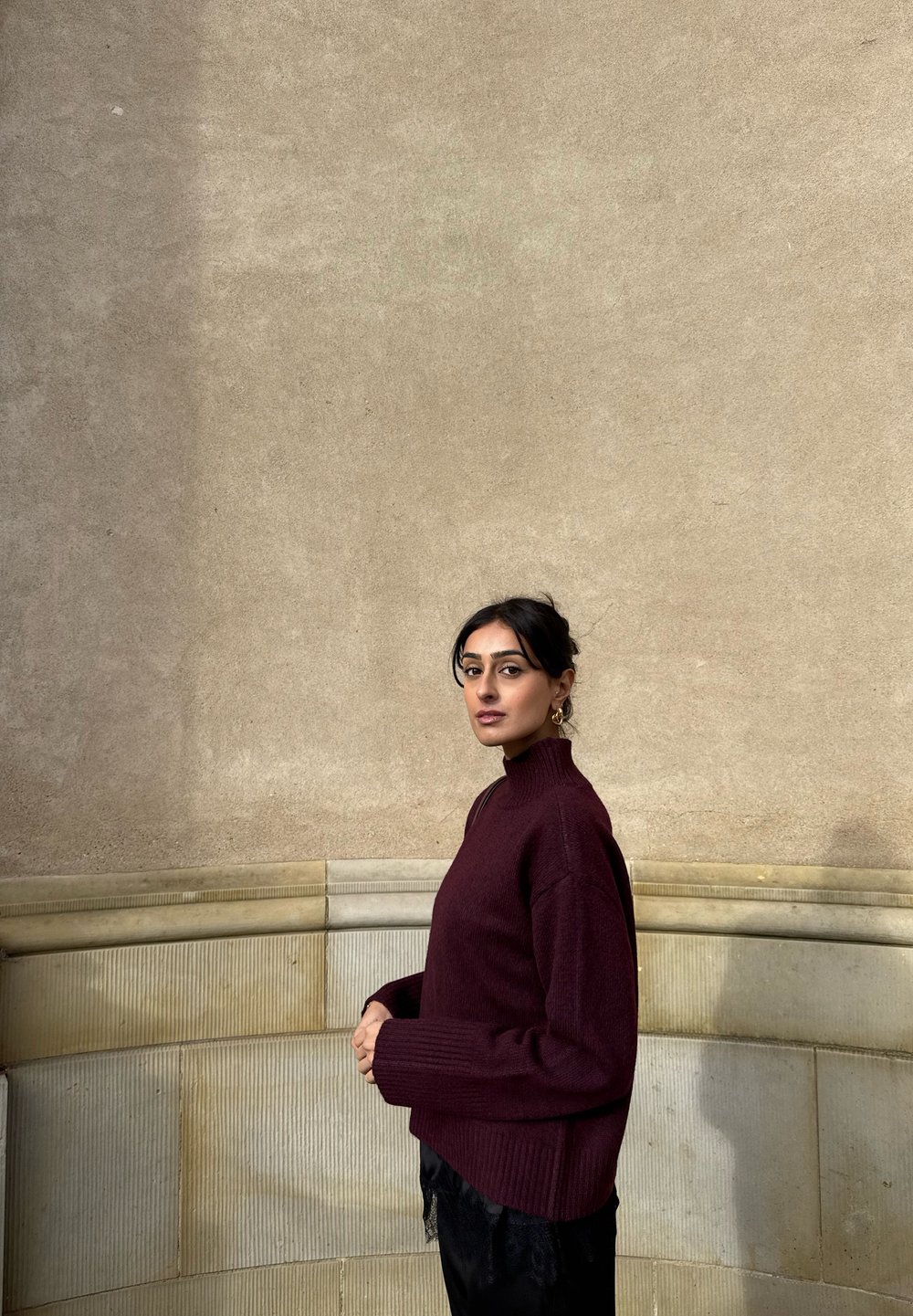 Bordeauxrode geribbelde coltrui met een losse pasvorm, gecombineerd met zwarte, gestructureerde broek. Soepele, zachte textuur. Gefotografeerd tegen een lichte stenen muur.