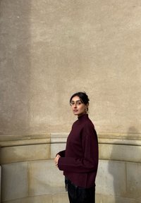 Bordeauxrode geribbelde coltrui met een losse pasvorm, gecombineerd met zwarte, gestructureerde broek. Soepele, zachte textuur. Gefotografeerd tegen een lichte stenen muur.