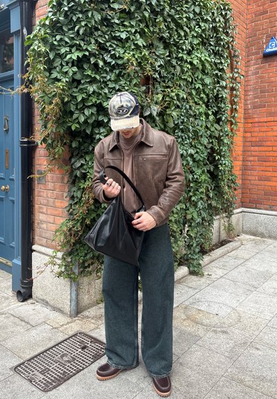 Chaqueta de cuero marrón sobre una camisa marrón, pantalones de mezclilla verdes anchos, sosteniendo un bolso de cuero negro, con una gorra de camuflaje. Follaje verde de fondo.