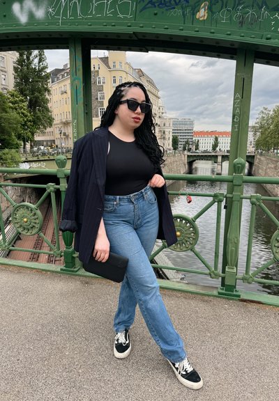 Bodysuit negro, chaqueta de rayas, jeans de tiro alto azules, clutch negro, zapatillas negras con suela blanca y gafas de sol, posando junto a un puente verde.