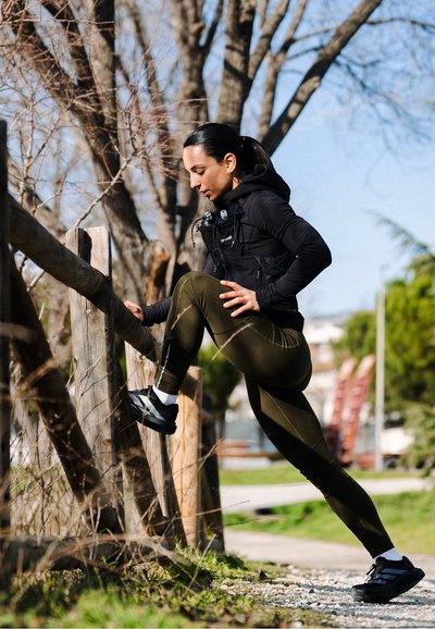 Mujer con ropa deportiva estirando la pierna en una cerca de madera al aire libre, con árboles y un camino bajo un cielo despejado.