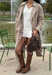 Donna che indossa un trench beige, un maglione bianco a maglia, pantaloni corti bianchi e stivali marroni fino al ginocchio, mentre tiene una borsa marrone vicino a una fontana e sedie verdi.