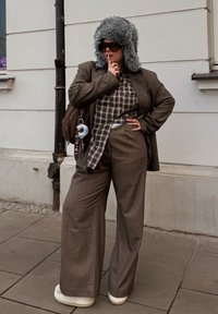Costume marron avec pantalon à jambes larges et blazer long assorti porté sur une chemise à carreaux. Chapeau gris doublé de fourrure et chaussures blanches complètent le look.