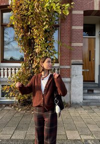 Bruine gebreide cardigan met grote knopen, witte blouse eronder, gecombineerd met geruite broek. Zwarte tas met bedeltje; achtergrond van bladeren en bakstenen.