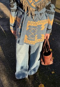 Pull enveloppant bleu à motifs géométriques, bords frangés, associé à un jean denim clair à jambes larges et à un sac à main en cuir marron. Chaussures noires.