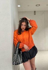 Oranžová bunda na zips s elastickými manžetami, čierna volánová sukňa a čierna taška so striebornými cvočkami. Hladké textúry, moderný dizajn.