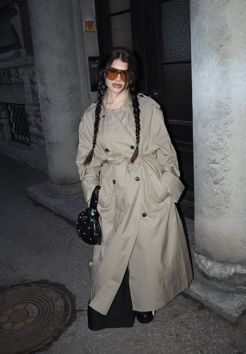 Kvinde med lange fletninger iført beige trenchcoat og ravfarvede solbriller, holder en sort nittebesat taske, stående mellem stenskulpturer udendørs om natten.