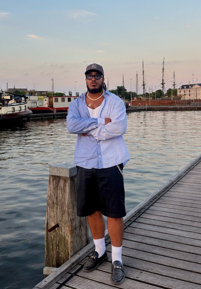 Un hombre se encuentra en un muelle de madera junto al agua, vistiendo una camisa de rayas azul claro, pantalones cortos negros, calcetines blancos y mocasines negros. Se pueden ver detalles de cadenas plateadas.