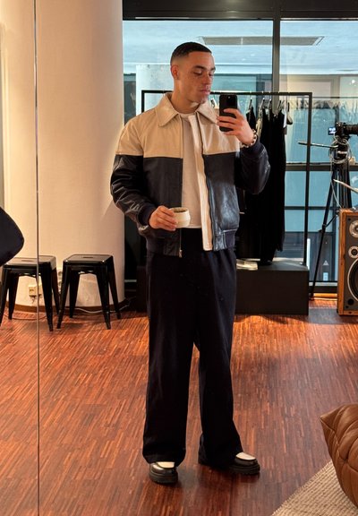 Chaqueta bomber bicolor con una parte superior crema y una inferior azul marino, combinada con pantalones oscuros y zapatos negros gruesos. Sostenido un vaso.