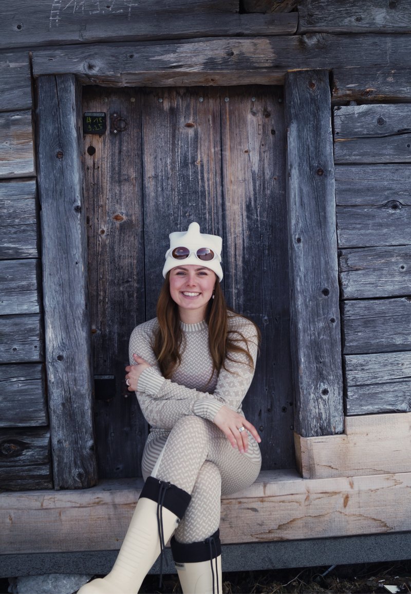 Femme souriante en maille beige, chapeau blanc et lunettes de soleil assise sur un banc en bois devant une porte de cabine en bois sombre usée.