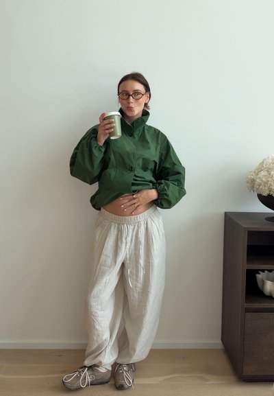 Mujer con gafas y ropa holgada sostiene una taza verde cerca de su boca mientras toca su cintura, de pie junto a un armario de madera oscura con flores.