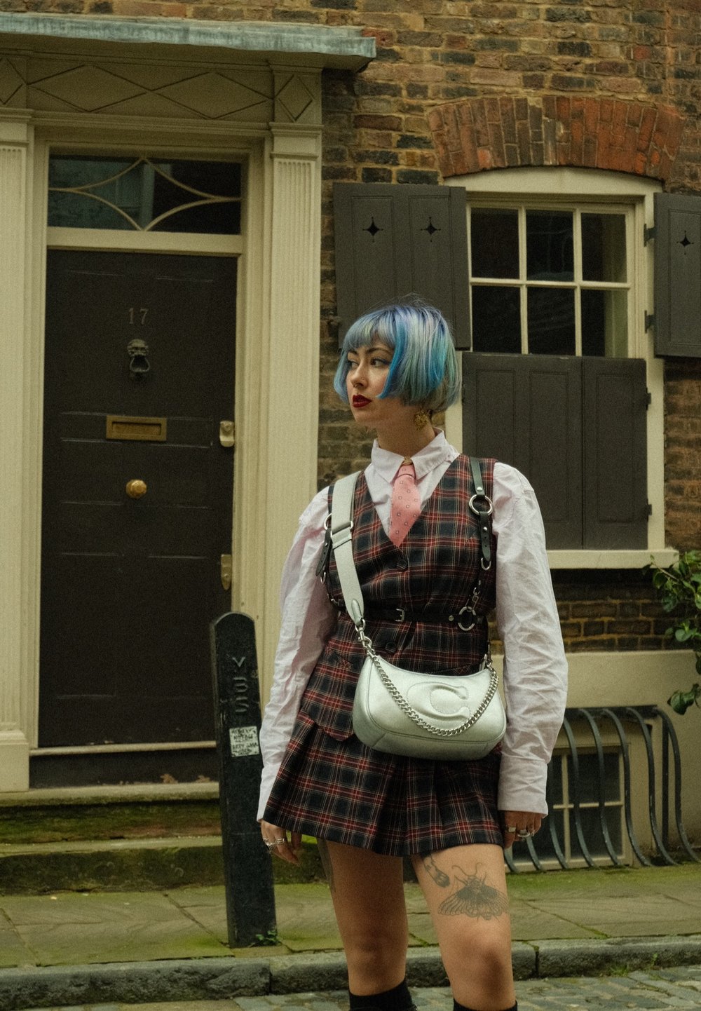 Καρό φόρεμα με σκούρες και ανοιχτές αποχρώσεις, λευκό πουκάμισο, ροζ γραβάτα; ασημένια τσάντα με αλυσίδα; κοντά μαλλιά με μπλε ανταύγειες και τατουάζ στο πόδι.