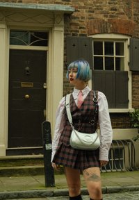 Καρό φόρεμα με σκούρες και ανοιχτές αποχρώσεις, λευκό πουκάμισο, ροζ γραβάτα; ασημένια τσάντα με αλυσίδα; κοντά μαλλιά με μπλε ανταύγειες και τατουάζ στο πόδι.