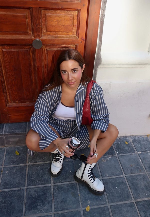 Gestreiftes Stoffoutfit mit lockerer Passform, bestehend aus einem Button-Up-Hemd und Shorts. Weiße Combat-Stiefel, hält Sonnenbrille und ein Getränk. Hintergrund aus Holztür.