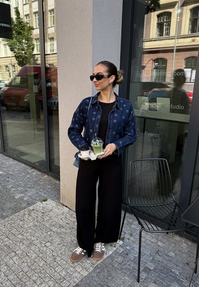 Mujer con gafas de sol, vistiendo una chaqueta de mezclilla con patrón de corazones y pantalones negros, sosteniendo una bandeja con una taza de café y un batido verde fuera de un café.