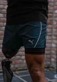 Sinakas spordipüksid valgete aktsentidega, millel on Puma logo. Kandmisel mustade surupükstega, mis on kombineeritud proteesijalaga.